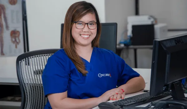 female medical office adminstrator in blue scrubs sits at computer and smiles for camera