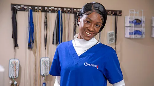 A Neurodiagnostic Technologist in scrubs smiles as she stands in front of health diagnostic equipment..