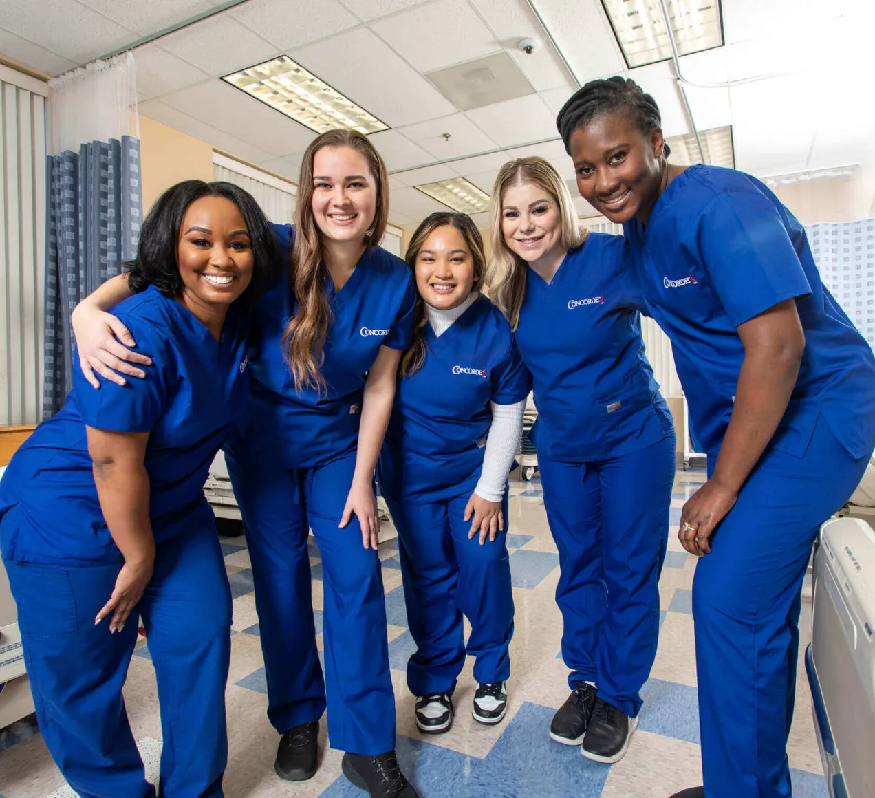 5 smiling Concorde graduates in blue scrubs in lab setting