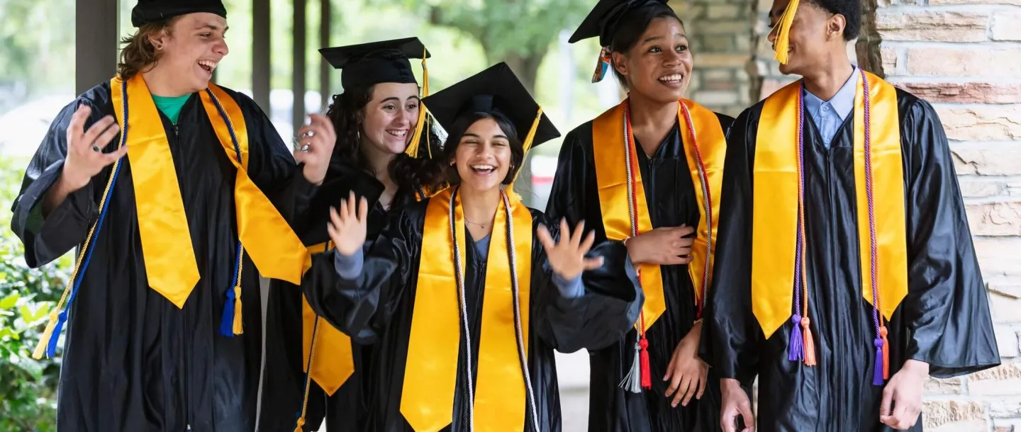 Five diverse high school seniors wearing black graduation caps and gowns with gold stoles smile and celebrate together, representing recipients of a high school senior grant.