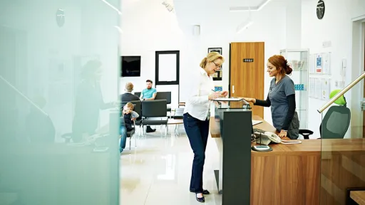 A young medical assistant in scrubs stands behind a desk and interacts with a patient in a waiting room at a medical facility.