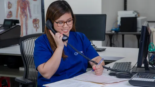 A medical office administrator sits in front of her computer and answers a phone call from an unseen patient.