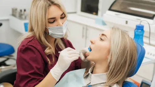Dental hygienist in maroon scrubs using a dental mirror to examine patient’s mouth during checkup