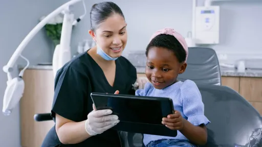 A female dental assistant in black scrubs and medical gloves talking to child patient and showing patient a tablet dental assistant responsibilities