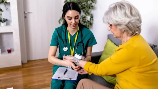 A young female medical assistant in green scrubs uses a digital cuff to take the blood pressure of an older female patient while reviewing paperwork on a clipboard.