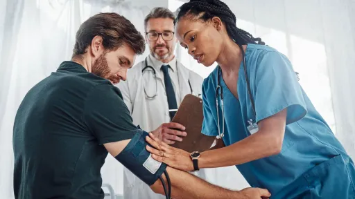 A medical assistant in blue scrubs carefully applies a blood pressure cuff to a patient's arm while a doctor stands behind them holding a clipboard and observing.