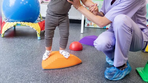 A pediatric physical therapist assistant in purple scrubs provides manual support to a young child practicing standing and balance on a therapeutic platform.