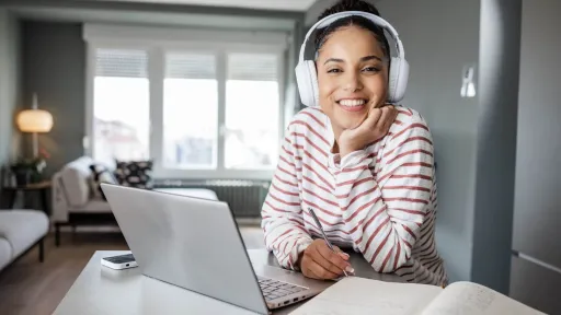 A smiling female student wearing white headphones sits at a desk with a laptop and notebook, studying for an admission test in a bright home office.