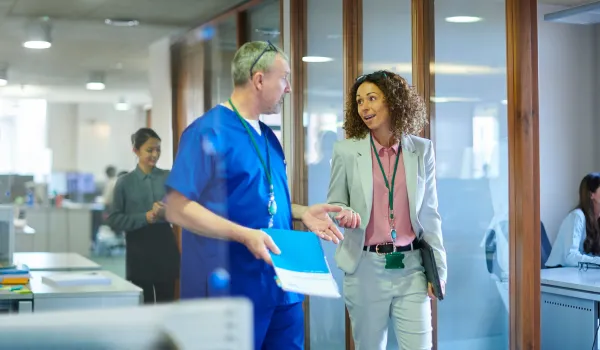 A healthcare administrator speaks with a medical provider as they walk together down the hallway of a medical facility.