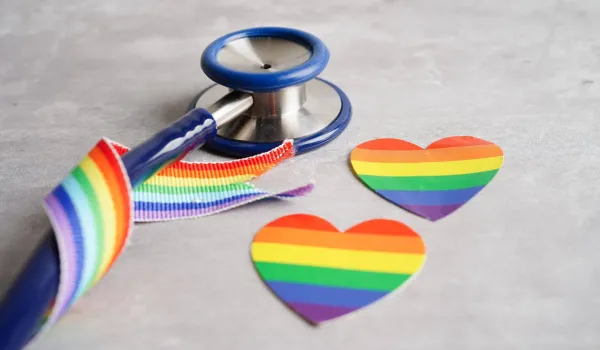 Stethoscope wrapped around rainbow pride ribbon with rainbow pride flag and pride flag hear cutouts on table