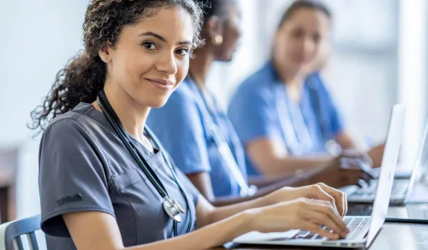 Smiling medical assistant student with stethoscope using laptop in class, representing medical assistant certification training.
