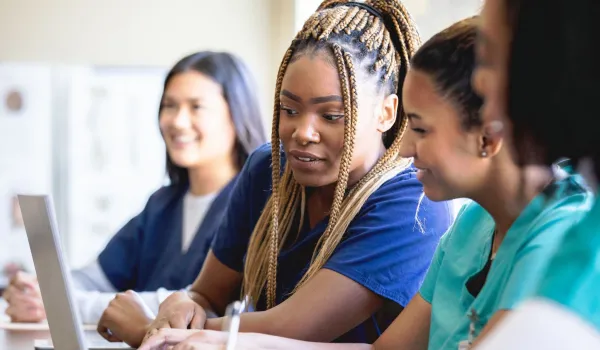 A diverse group of female healthcare career training students in scrubs are gathered around a laptop in a bright classroom setting, looking at the screen together.