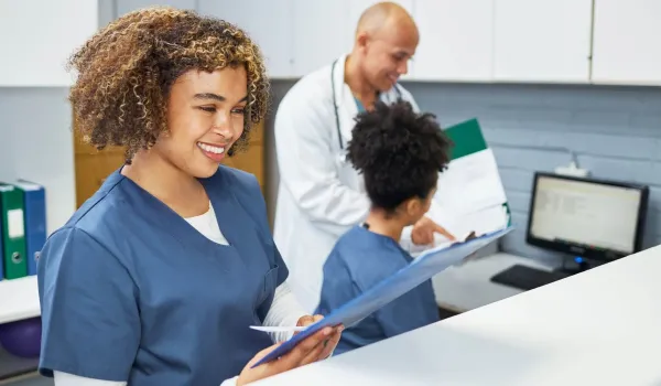 A smiling medical assistant in blue scrubs stands at a reception counter reviewing a patient chart while a doctor and another medical assistant discuss data on a computer screen in the background.
