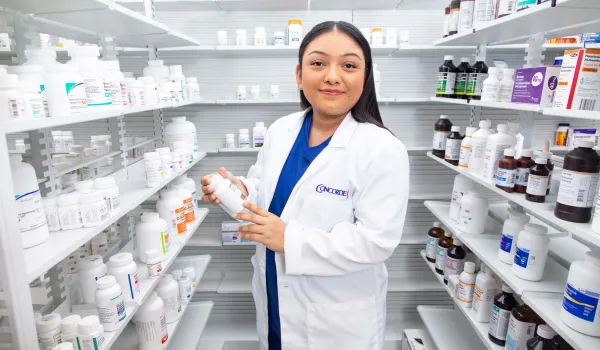 A smiling female pharmacy technician in a white lab coat stands in an aisle between shelves full of medication, holding a bottle to check the label.