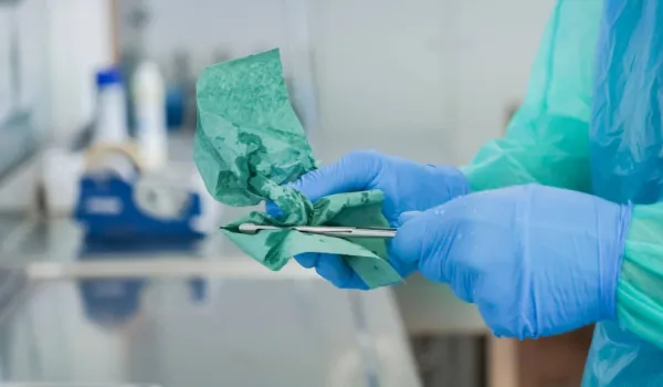 A person wearing a blue protective gown and surgical gloves carefully wipes and cleans a small surgical instrument during the decontamination phase of sterile processing.