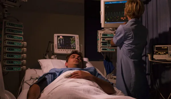 A sleep technologist monitors real-time physiological data and vital signs on multiple screens while a patient sleeps in a darkened room.
