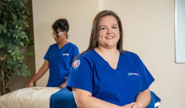 A smiling Concorde massage therapy student wearing blue scrubs with a military-themed patch stands in a healthcare training lab, illustrating how to use military tuition assistance for career training.