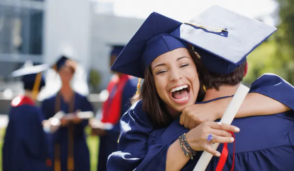 Female graduate in blue cap and gown holding diploma hugs another graduate with big smile on her face.
