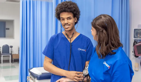 A male medical assistant wearing blue scrubs and stethescope works with patient and smiles for camera