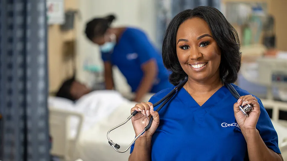 LPN in scrubs smiles while holding her stethoscope.