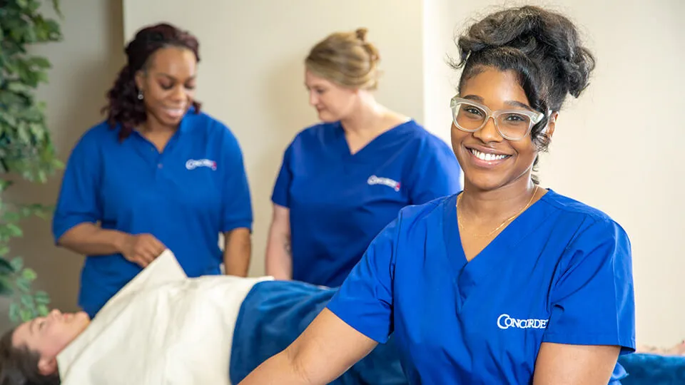 A massage therapist leans on her table and smiles at the camera.