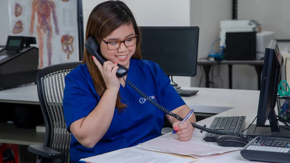 A medical office administrator sits in front of her computer and answers a phone call from an unseen patient.