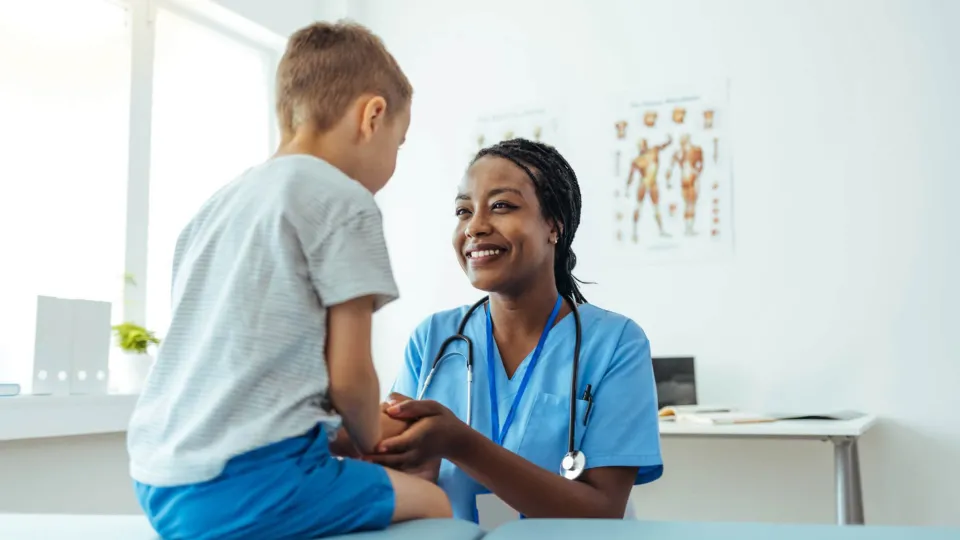 Smiling medical assistant supports young patient during checkup in clinical exam room setting.