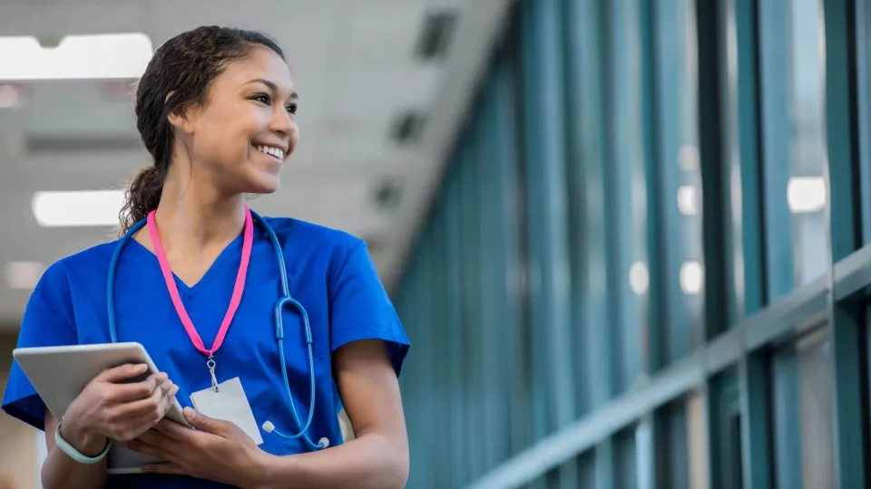 Smiling travel nurse in scrubs holding a tablet in a hospital hallway, representing confidence and readiness for a traveling nurse career