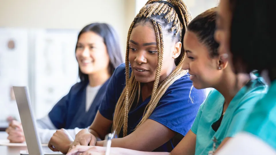 A diverse group of female healthcare career training students in scrubs are gathered around a laptop in a bright classroom setting, looking at the screen together.