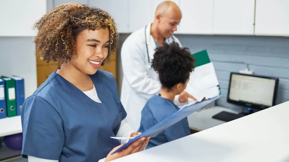 A smiling medical assistant in blue scrubs stands at a reception counter reviewing a patient chart while a doctor and another medical assistant discuss data on a computer screen in the background.