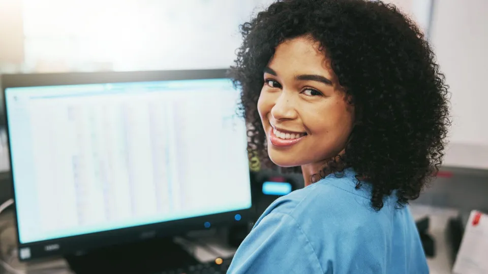 Smiling woman in blue scrubs working at a computer, representing Medical Office Administration Certificate Program skills.
