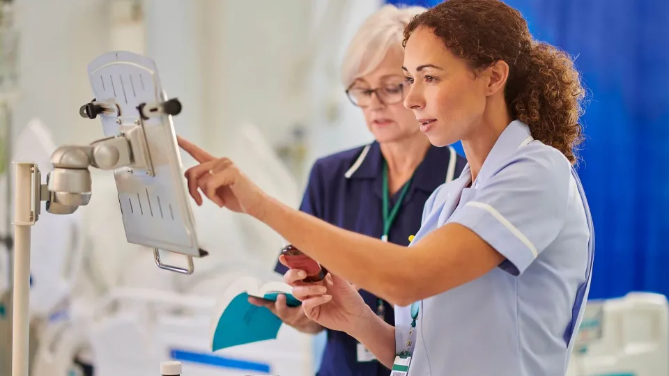 Two female nurses in professional uniforms review medical data on a wall-mounted digital terminal while one holds a medication bottle and a reference book.