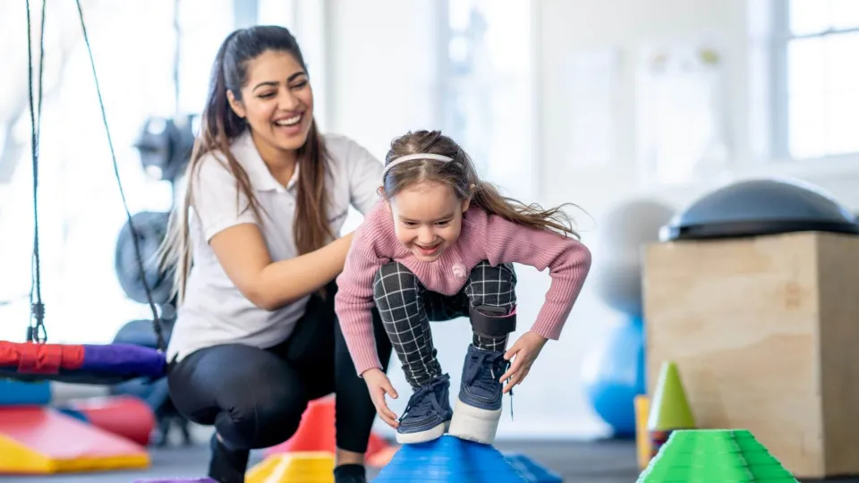 A young child balances on a large blue therapy ball while being guided through stability exercises by a physical therapist assistant.