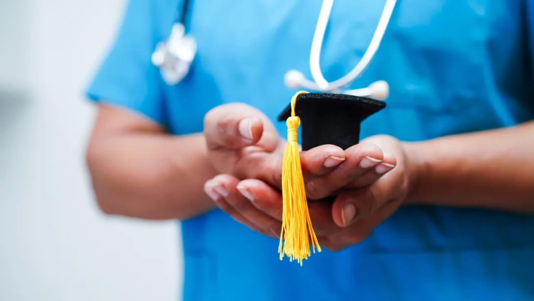 A medical professional wearing scrubs and a stethoscope holds a miniature graduation cap.